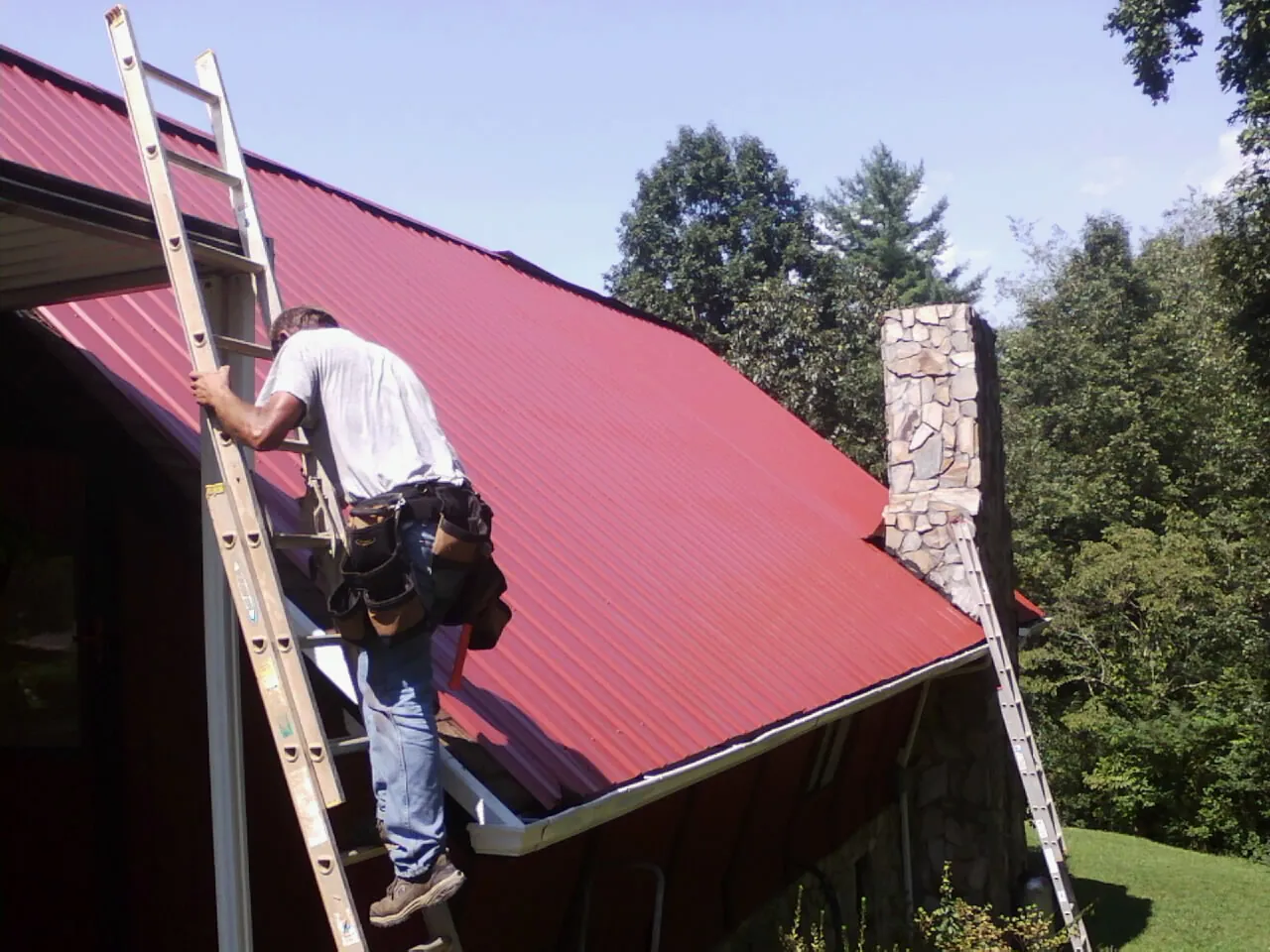 Roofer on a ladder beside a red metal roof and stone chimney — Soffit & Fascia Repair by Roofing Brothers in Box Springs